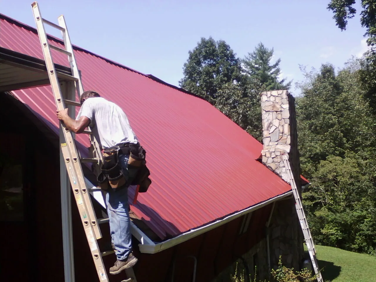 Roofer on a ladder beside a red metal roof and stone chimney — Soffit & Fascia Repair by Roofing Brothers in Bay Pines